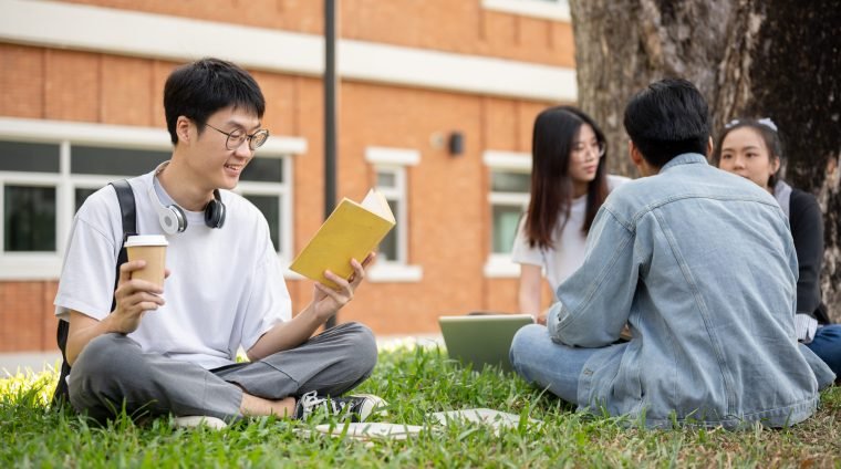 A positive and nerd Asian male college student is reading a book in a campus park with his friends. A positive and nerd young Asian male college student is reading a book while relaxing sitting on grass in a campus park with his friends. Education concept